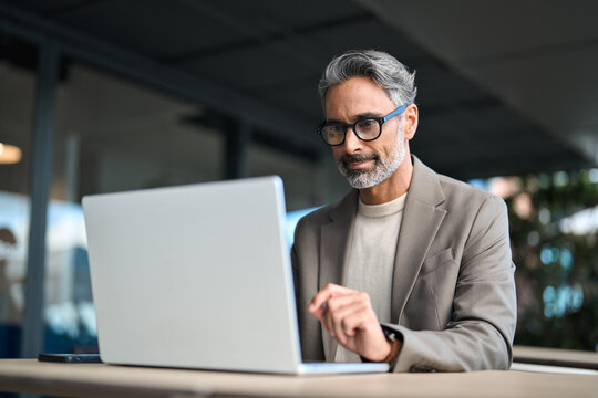 Busy middle aged mature professional business man entrepreneur wearing suit and eyeglasses sitting at table outdoors working online using laptop computer hybrid working outside office.
