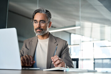 Mature professional businessman manager working looking at computer technology in office. Busy middle aged Latin business man executive ceo wearing suit and glasses sitting at desk using laptop.