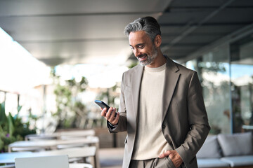 Smiling handsome middle aged professional businessman holding cellphone looking at smartphone standing outside. Happy mature business man executive using mobile cell phone outdoors. Candid photo