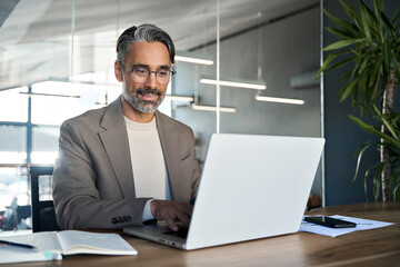 Mature professional businessman manager working looking at computer technology in office. Busy middle aged Latin business man executive ceo wearing suit and glasses sitting at desk using laptop.
