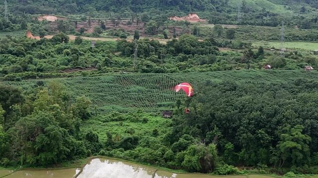 Drone circles above a paramotor approaching for landing in Vang Vieng, Laos, panning down to capture a top-down view over the grassy field