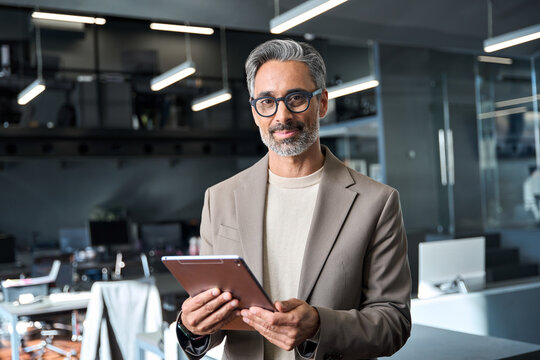 Portrait of happy mature older business man executive standing in office using digital tablet. Middle aged businessman manager wearing suit glasses working on professional financial project.