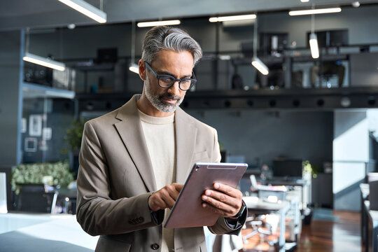 Mature older businessman professional executive manager investor working on tech tab device. Busy middle aged business man ceo wearing suit standing in corporate office using digital tablet.
