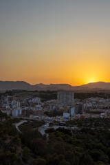 Obraz premium Aerial view of Alicante city at sunset, showcasing the skyline against a vibrant orange and blue sky, highlighting buildings and lush greenery in the foreground.