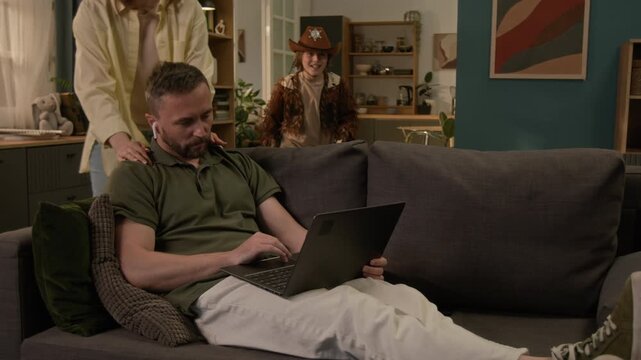 Woman speaking with husband touching his shoulders, then taking playful kids away as man asking for silence when working on laptop remotely sitting on couch