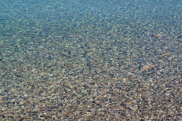 Full frame overhead shot of a clear, calm body of water revealing countless pebbles and stones on the bottom, with sunlight creating gentle ripples and shimmering patterns on the surface