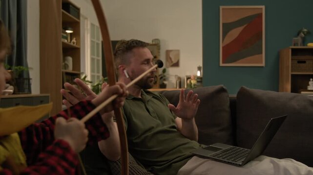 Younger child aiming toy bow at elder brother who popping up behind couch making faces, their father working on laptop and asking kids to stop playing - Powered by Adobe