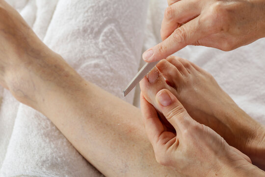 Elderly woman taking care of herself by doing her own pedicure of damaged feet and broken toe nails at home. Close-up of hands during the beauty routine. Natural light and cozy home spa. 