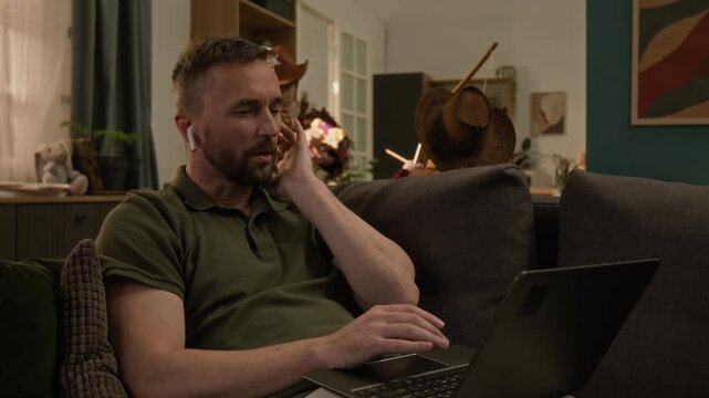 Man with earbuds and laptop on his lap communicating online while working from home, two kids in costumes playing in background, woman taking them away