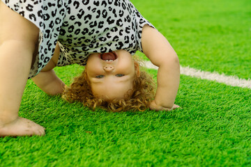A young girl with curly hair enjoys a playful moment outside doing a handstand on bright green...