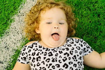 A young girl with curly hair is lying on green grass playfully sticking out her tongue She wears a...