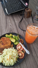 Vertical photo of healthy food, while working, near a laptop, lunch at the office, with a delicious melon water with ice, breaded chicken with white rice, and vegetables.