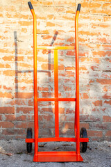 A bright orange hand truck sits against a brick wall, ready to move heavy objects
