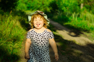 A cheerful child stands on a grassy pathway wearing a playful outfit and hat joyfully sticking out...