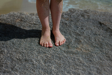 Child feet covered in golden sand standing on weathered limestone rocks at Mediterranean beach