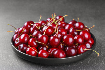 Close up of black plate filled with ripe red sweet cherry placed on dark gray textured background. Delicious food, juicy summer fruit and healthy eating.