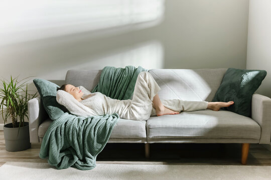 Woman in soft loungewear resting on a modern sofa with green blankets and pillows.