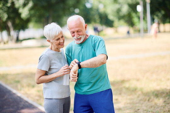 Smiling active senior couple jogging exercising and having fun and laughing together taking a break in the park, senior athletes checking their er heart rate with smartwatch - Powered by Adobe