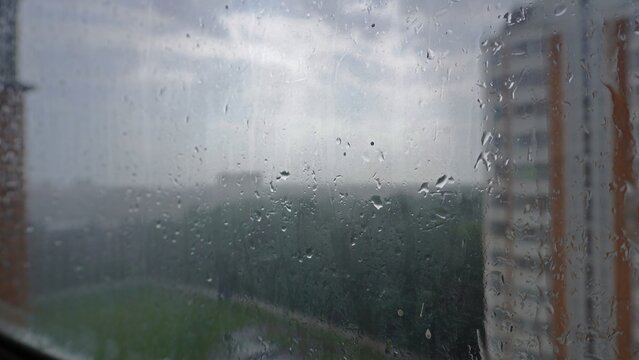 Fototapeta View from the window of the city buildings and trees during the rain. Water droplets are visible on the glass, creating a blurry and atmospheric effect.