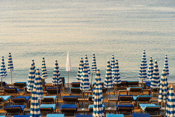 Rows of closed blue and white striped beach umbrellas and loungers on a tranquil pebble beach in Nice, French Riviera, during a calm sunrise. © Dmitri