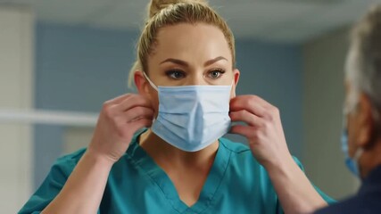 Woman in scrubs putting on a surgical mask in front of another person with a mask on face