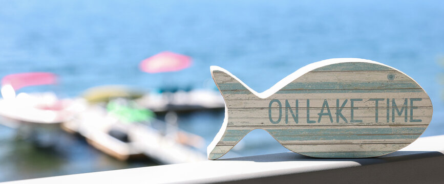 A small fish shaped wooden sign that says On Lake Time outside on a hot summer day near the water. 