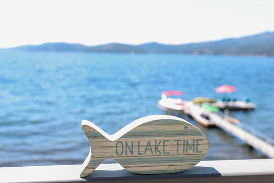 A small fish shaped wooden sign that says On Lake Time outside on a hot summer day near the water. 