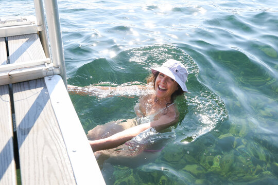Woman having fun in the lake. She is laughing and smiling while wearing a cute bucket hat. She is climbing out of the water on a dock ladder. 