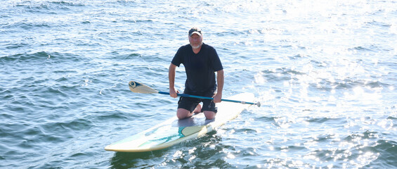 Happy middle aged man resting on his knees on a paddle board outside on a lake. He is holding a paddle over the water and working to maintain balance. 
