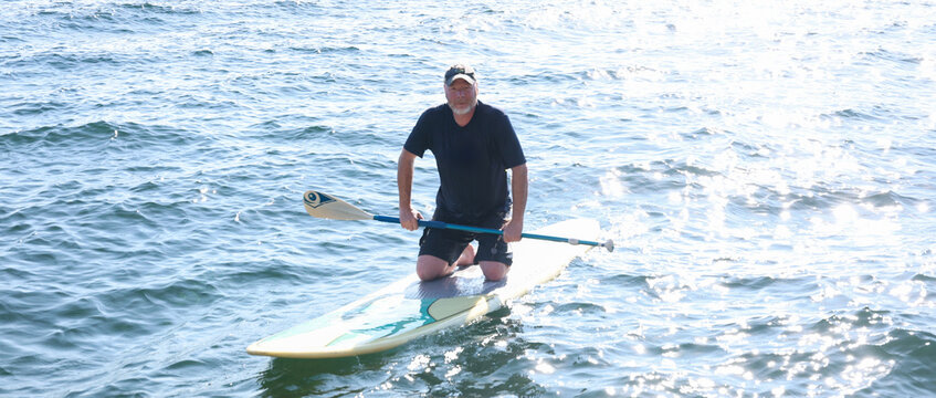 Happy middle aged man resting on his knees on a paddle board outside on a lake. He is holding a paddle over the water and working to maintain balance. 