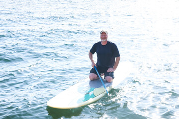 Happy middle aged man resting on his knees on a paddle board outside on a lake. He is holding a paddle over the water and working to maintain balance. 