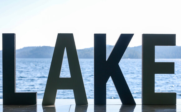 A sign with multiple letters forming the word Lake. Water from coeur D'Alene can be seen in the background