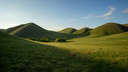 Rolling green hills under a clear blue sky, with gentle slopes and lush grass, creating a serene and tranquil landscape for nature enthusiasts and outdoor lovers