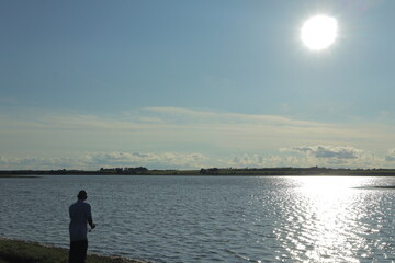 Fototapeta premium Peaceful Man Fishing at Lakeside During Golden Hour Sunset – Serene Countryside Landscape, Sparkling Water, and Clear Blue Sky in Summer Evening Tranquility