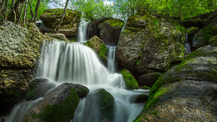 Cascading waterfall flows over moss-covered rocks surrounded by lush greenery, creating a serene...