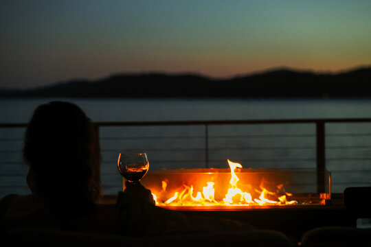 A woman sitting in an elegant setting at night outside drinking a glass of red wine with a warm fire overlooking a lake view. 