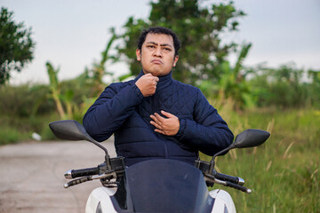 Asian man with a serious expression zips up his navy blue jacket while seated on a scooter on a rural path, preparing for a journey.