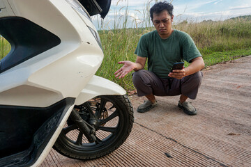 A frustrated man crouches by his broken-down scooter on a rural road, seeking help on his smartphone.