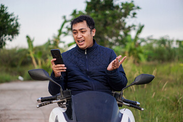 Excited Asian man on a motorcycle looking at his smartphone with a surprised expression, celebrating good news outdoors