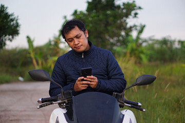 A young Asian man in a blue jacket sits on his scooter, intently using his smartphone in a rural outdoor setting.