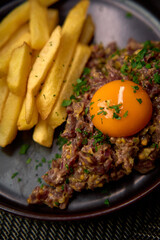 A delectable dish featuring a rich meat tartare topped with a vibrant egg yolk, accompanied by crispy golden fries and fresh herbs on a stylish plate.
