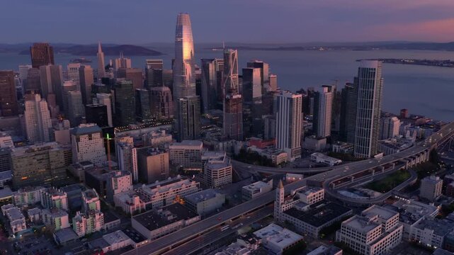 Aerial view of San Francisco, California, USA, at dusk. Skyscrapers dominate the skyline, reflecting the setting sun. Cars travel on the highway below.