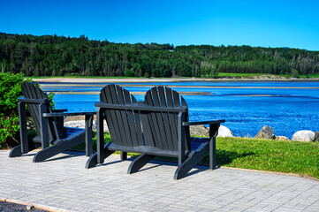 Adirondack chairs overlooking the serene river in La Malbaie, Quebec, with lush forest and bright blue water. Ideal image for concepts of relaxation, travel, and peaceful nature getaways.
