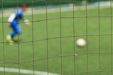 A soccer goal net in sharp focus, with a blurred background showing a player in a blue uniform about to kick the ball on a green artificial turf field