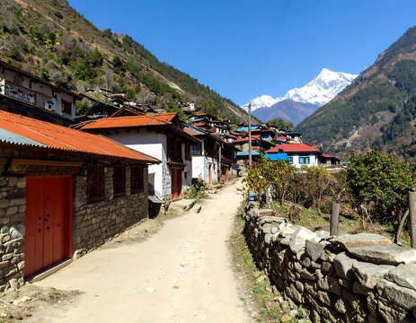 Mountain village nestled in a valley.  A dirt path winds through a cluster of traditional homes,  with colorful roofs and stone walls.  Snow-capped peaks in the distance