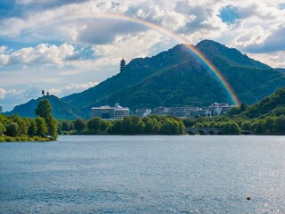 Rainbow arches over a serene lake with lush mountains and modern buildings in the background.