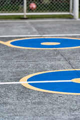 A playground soccer or basketball court featuring large blue circles outlined in yellow on a grey concrete surface. In the background, a blurry red ball is inside the goal