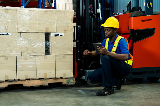 African male warehouse staff sitting on floor near boxes using tablet for checking stock data beside forklift inside storage facility, inside logistics warehouse.