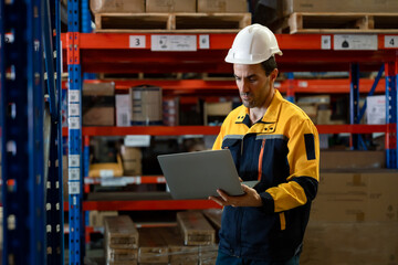 Warehouse worker white adult male using laptop while managing inventory between tall racks filled...
