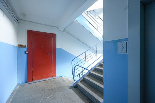 well-lit hallway with a red door on the left and stairs with blue railings on the right. An elevator call button is on the wall - Powered by Adobe